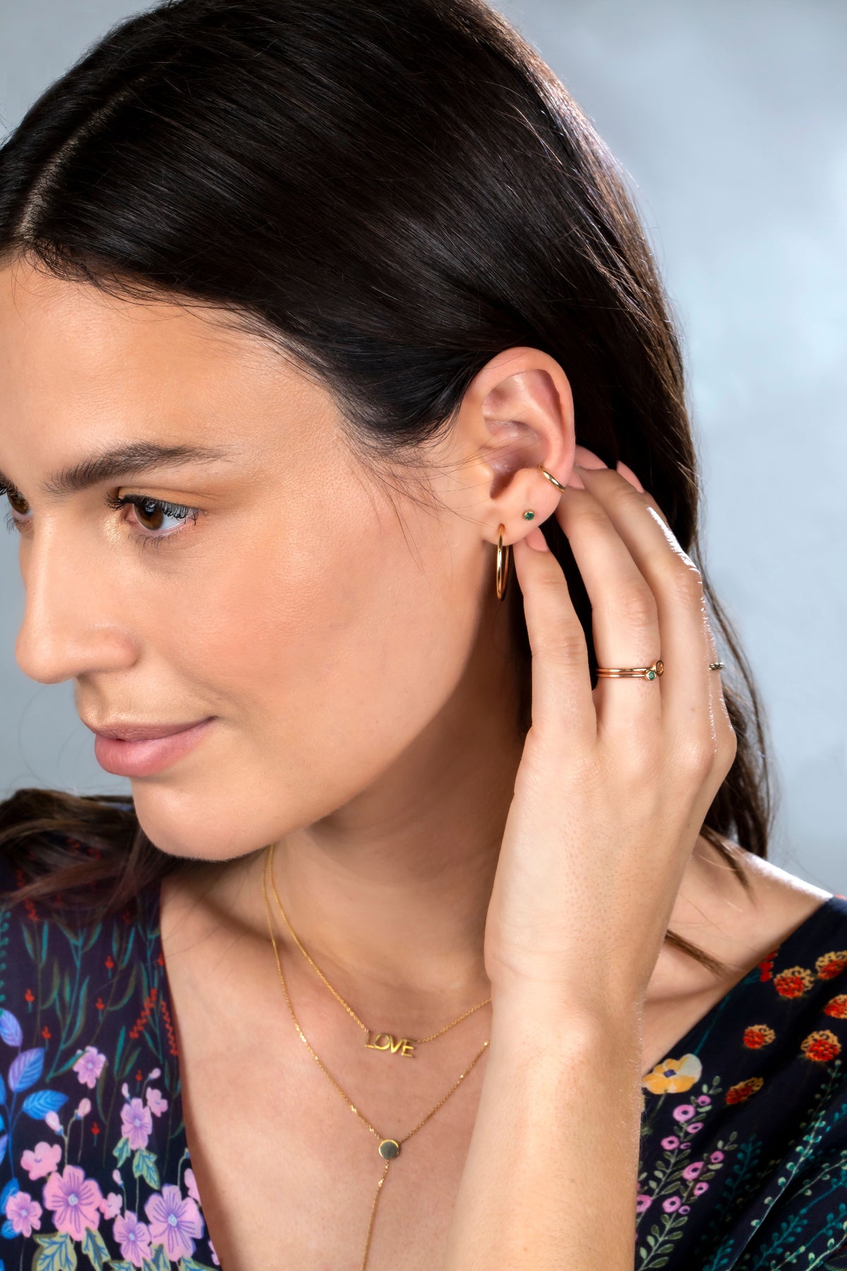 A Birthstone Stud, Rounded Ear Cuff, yellow gold hoop earring, two Birthstone Rings and LOVE Necklace worn by a brunette woman with a floral navy top against a light blue background. 