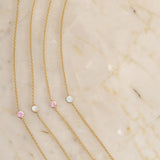 Four yellow gold 14K Gold Asymmetrical Birthstone Necklaces (two Opal and two Pink Tourmaline) on a light beige marble background. 