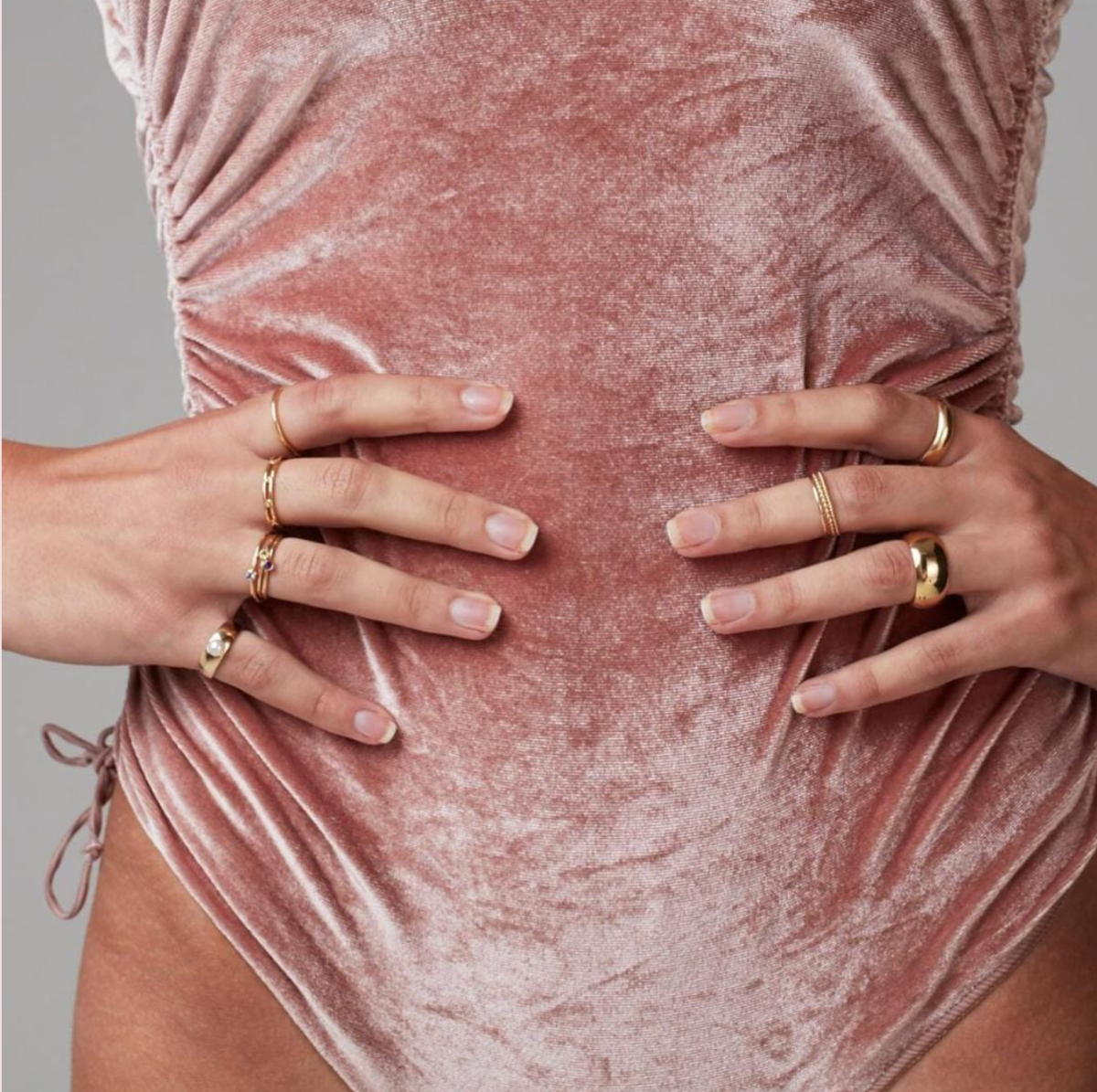 A close-up of hands on hips wearing a Pinky Perla Ring, three Birthstone Rings, a Link Ring, a Delicate Band, Delicate Trio Stacking Rings, a Thick Band and a Tube Ring, all in yellow gold, styled in a pink velvet leotard.