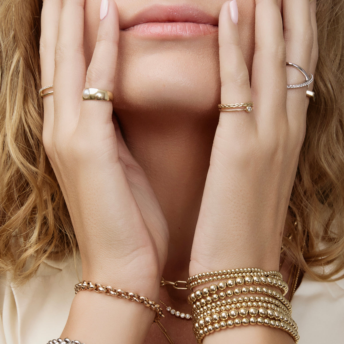 Close-up of a woman's hands wearing several yellow gold Bubble Bracelets, two Rolo Bracelets, a Perla Pinky Ring, Braided Ring, Birthstone Ring, Criss-Cross Diamond Duo Ring and Criss-Cross Diamond Daze Ring, placed on her face with her blonde hair and a cream shirt. 