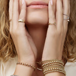 Close-up of a woman's hands wearing several yellow gold Bubble Bracelets, two Rolo Bracelets, a Perla Pinky Ring, Braided Ring, Birthstone Ring, Criss-Cross Diamond Duo Ring and Criss-Cross Diamond Daze Ring, placed on her face with her blonde hair and a cream shirt. 