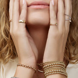 Close-up of a woman's hands wearing several yellow gold Bubble Bracelets, two Rolo Bracelets, a Perla Pinky Ring, Braided Ring, Birthstone Ring, Criss-Cross Diamond Duo Ring and Criss-Cross Diamond Daze Ring, placed on her face with her blonde hair and a cream shirt. 