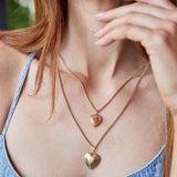 Close-up of a woman's décolletage wearing a Box Chain with a Heart Locket Charm and a Round Curb Chain with a Diamond Heart Locket Charm, all in yellow gold, wearing a denim blue singlet top with her orange hair visible.