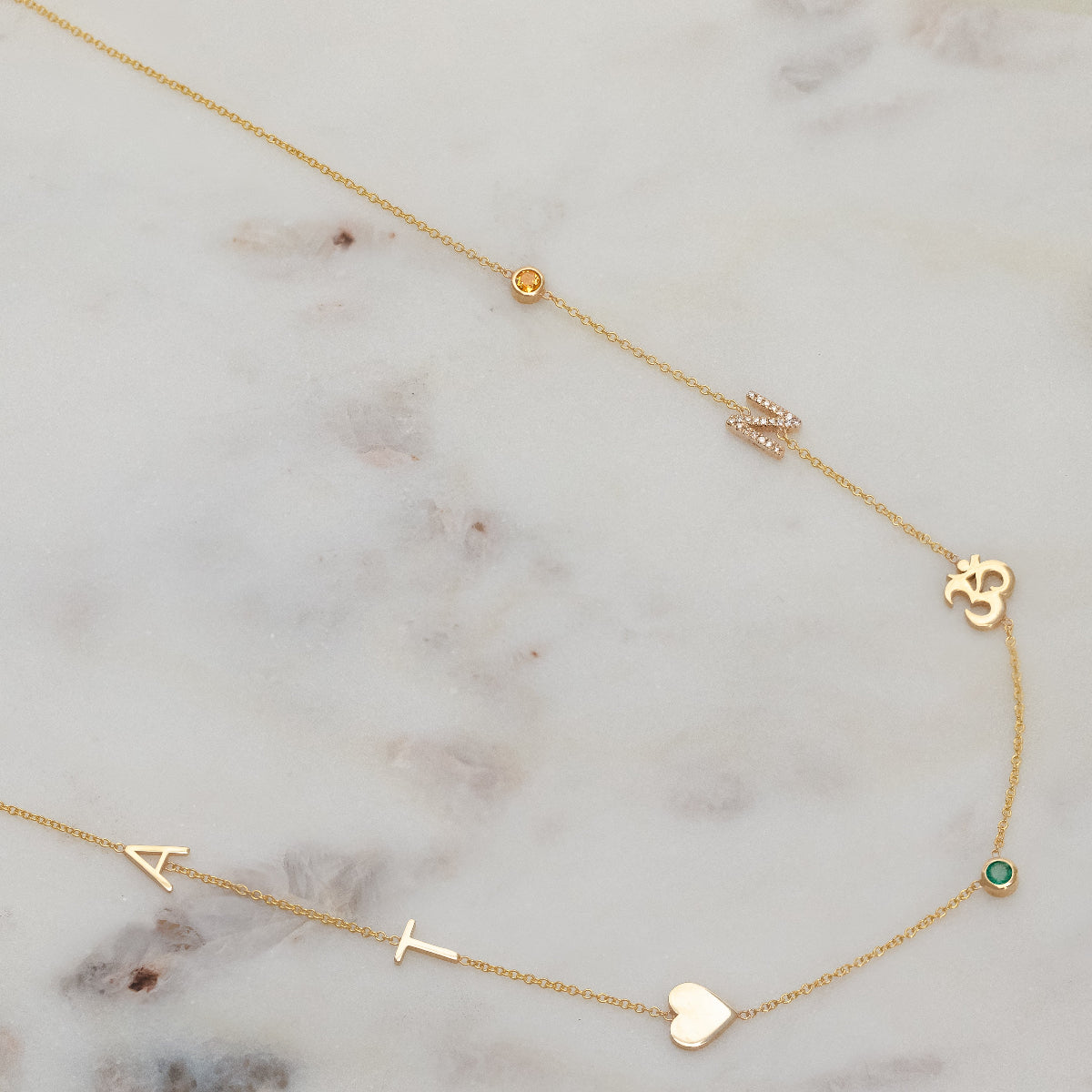 A Custom Gold Necklace in yellow gold with seven charms, the letters A and T, a heart, two birthstones, an om and a pavé letter N, on a marble surface.