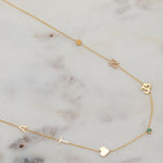 A Custom Gold Necklace in yellow gold with seven charms, the letters A and T, a heart, two birthstones, an om and a pavé letter N, on a marble surface.