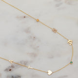 A Custom Gold Necklace in yellow gold with seven charms, the letters A and T, a heart, two birthstones, an om and a pavé letter N, on a marble surface.