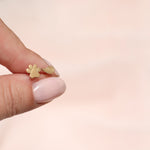 A hand holding a Charm Stud - Angel Wing and Paw in yellow gold against a light pink background. 