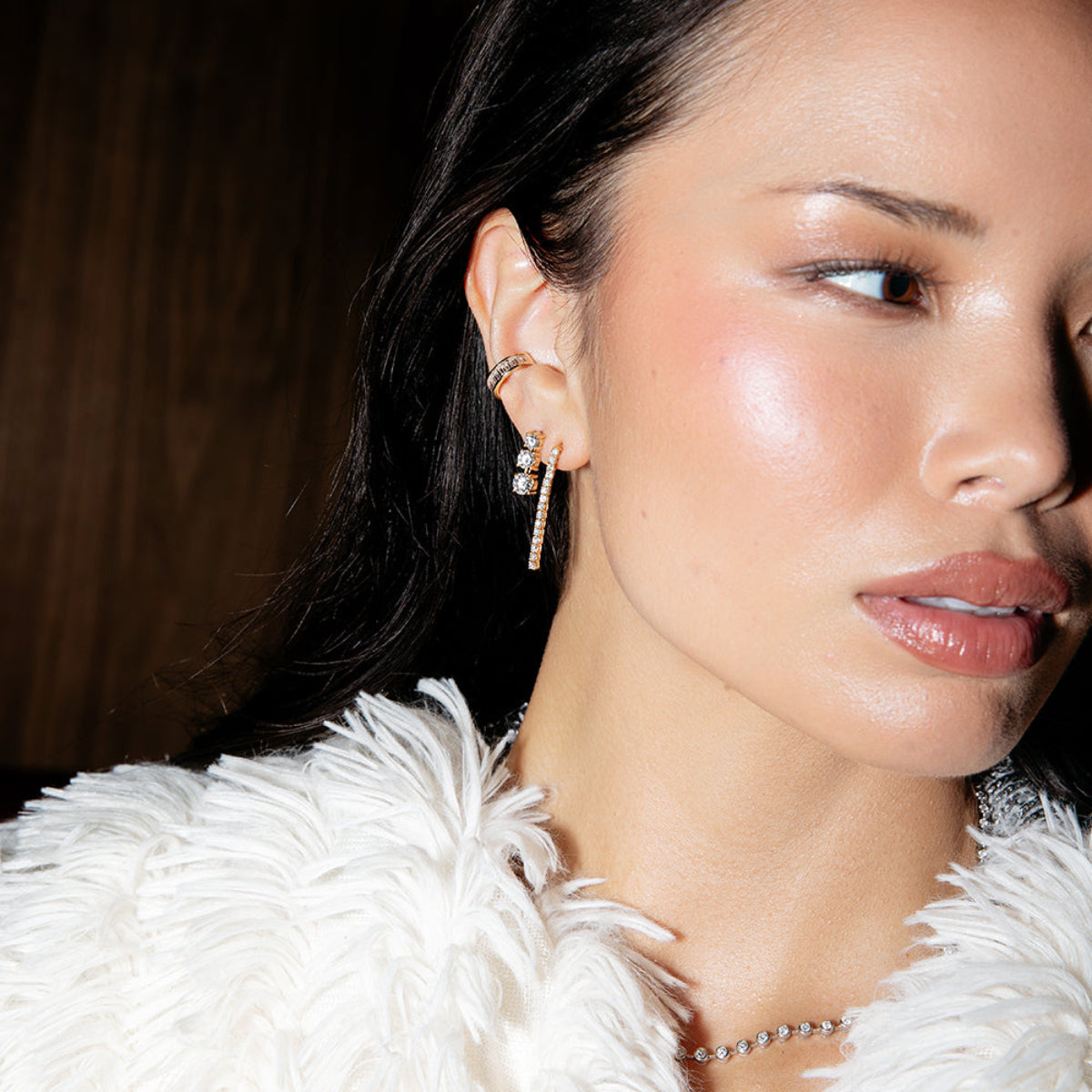 A brunette woman wearing the Diamond Cascade Earring, Triple Sec Diamond Earring, The Night Cap Ear Cuff and The Ultimate Diamond Necklace, styled with a cream fluffy jacket against a brown wooden background.