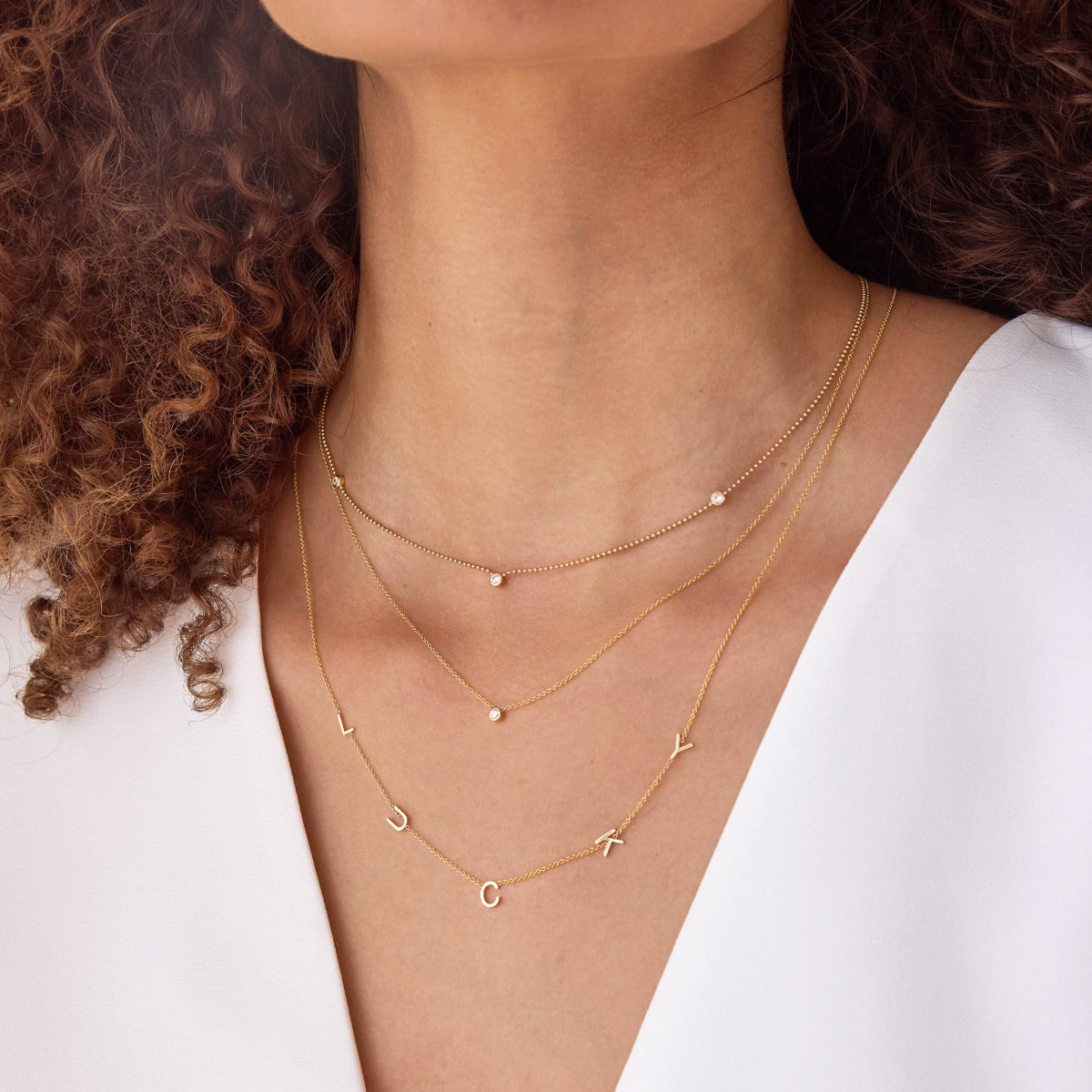 A close-up of a woman's neck wearing a Diamond Layering Necklace, a Triple Diamond Layering Necklace and a Custom Gold Necklace - 5 Letters, all in yellow gold, with brown curly hair and a white shirt.