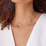 A close-up of a woman's neck wearing a Diamond Layering Necklace, a Triple Diamond Layering Necklace and a Custom Gold Necklace - 5 Letters, all in yellow gold, with brown curly hair and a white shirt.