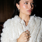 A woman wearing a Triple Sec Diamond Earring, The Night Cap Cuff, a Down the Line Earring, Make it a Double Ring, Cosmo Ring, Martini Ring, Pinky Promise Ring and The Ultimate Diamond Necklace, with a cream fluffy jacket against a dark background. 