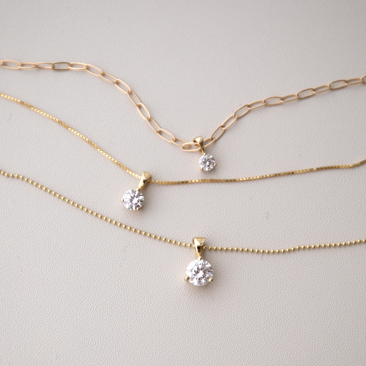 A Duchess Diamond Pendant on an Element Long Link Chain, a Princess Diamond Pendant on The Slide Chain and a Queen Diamond Pendant on a bead Chain in yellow gold against a light grey background. 