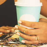A woman's hand wearing several rings, including a Geo Stacking Ring | Triangle and Petal Ring, with a black tank top holding a coffee cup. 