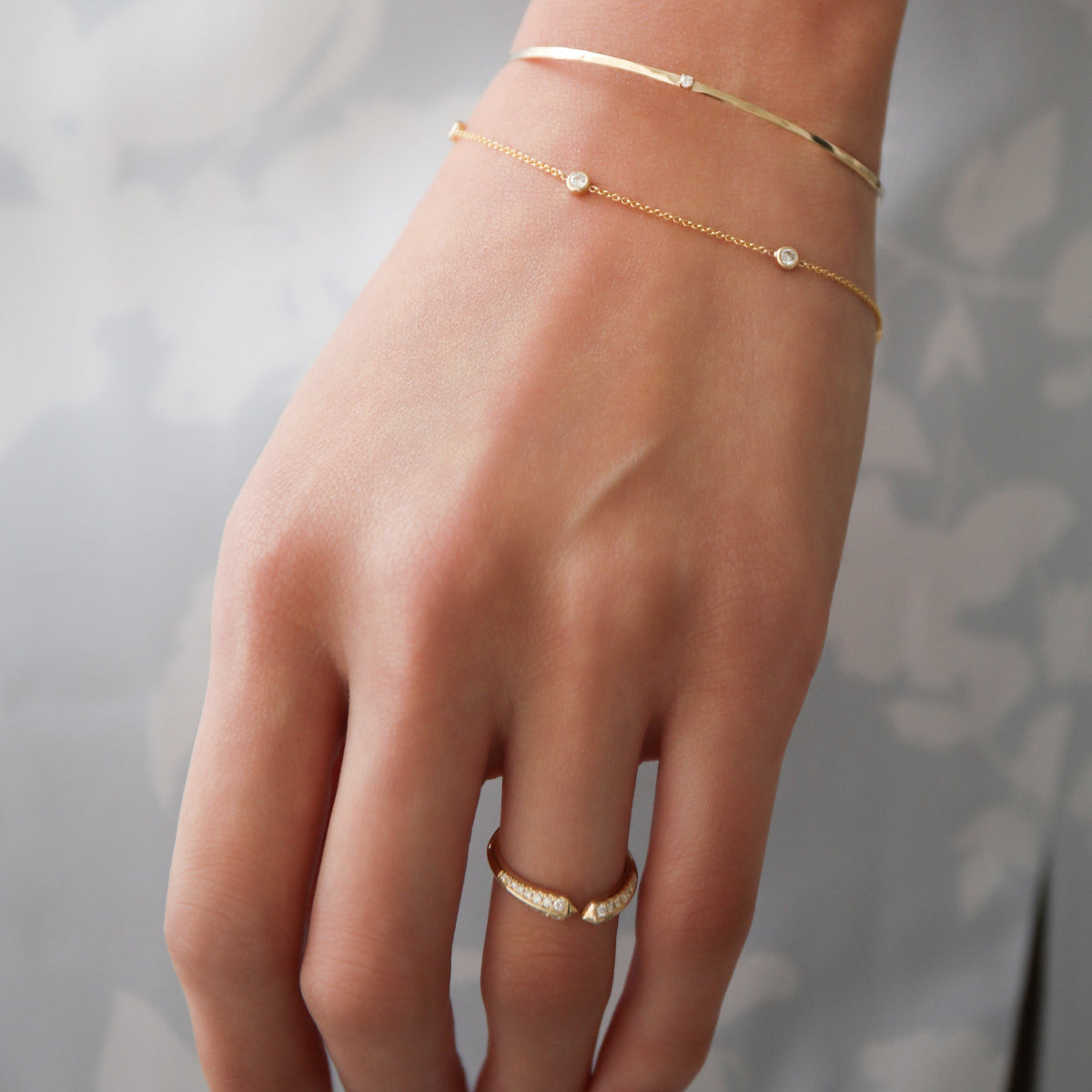 A close-up of a woman's hand wearing an Off the Cuff Bracelet, a Starstruck Diamond Bracelet and an Open Diamond Ring, against a white and light grey floral background.