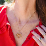 A close-up of a décolletage wearing a yellow gold Be Gentle in Grief Necklace with a red shirt and several rings. 