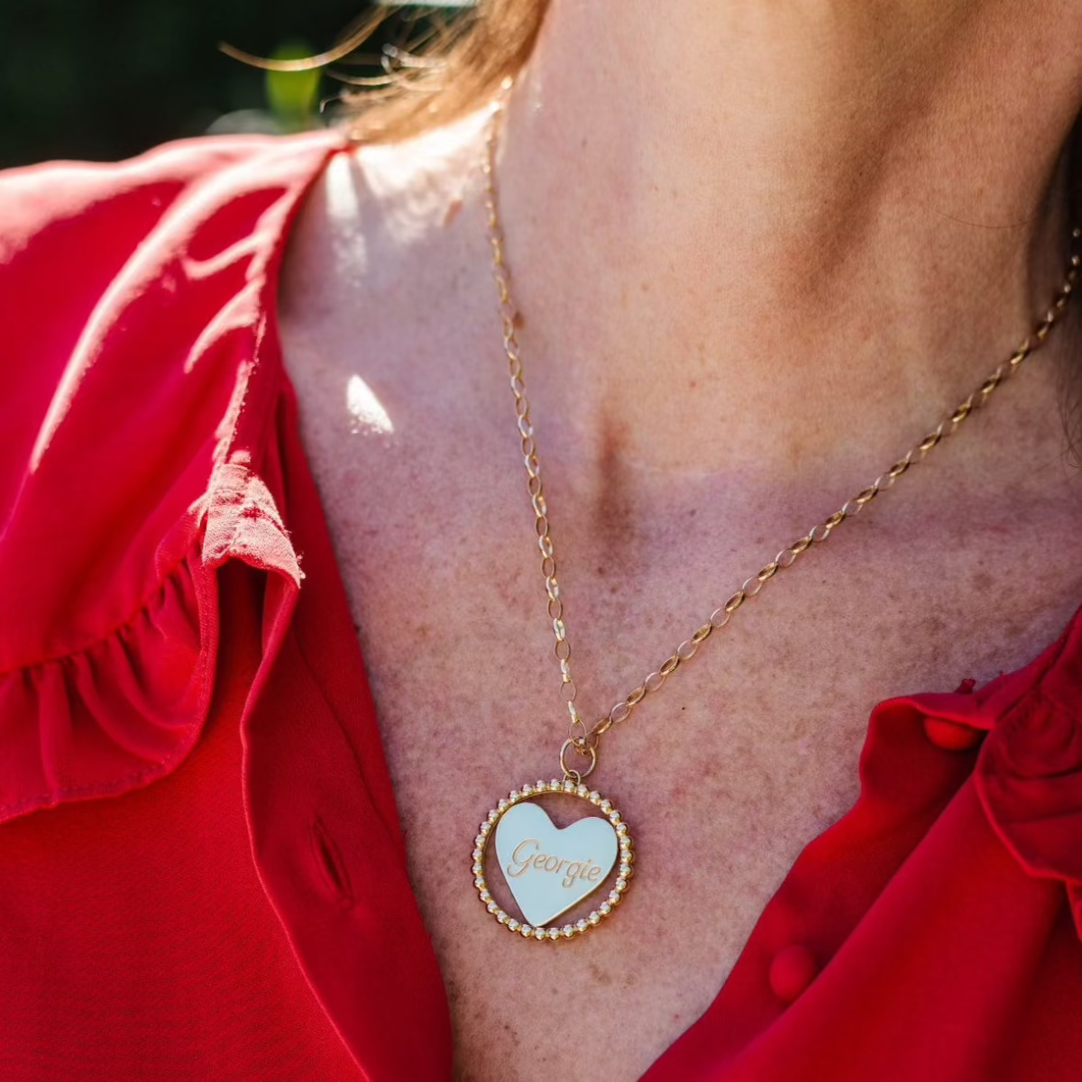 Close-up of a person's neck wearing a yellow gold Be Gentle in Grief Necklace engraved with 'Georgie,' paired with a red shirt and a blurred background.
