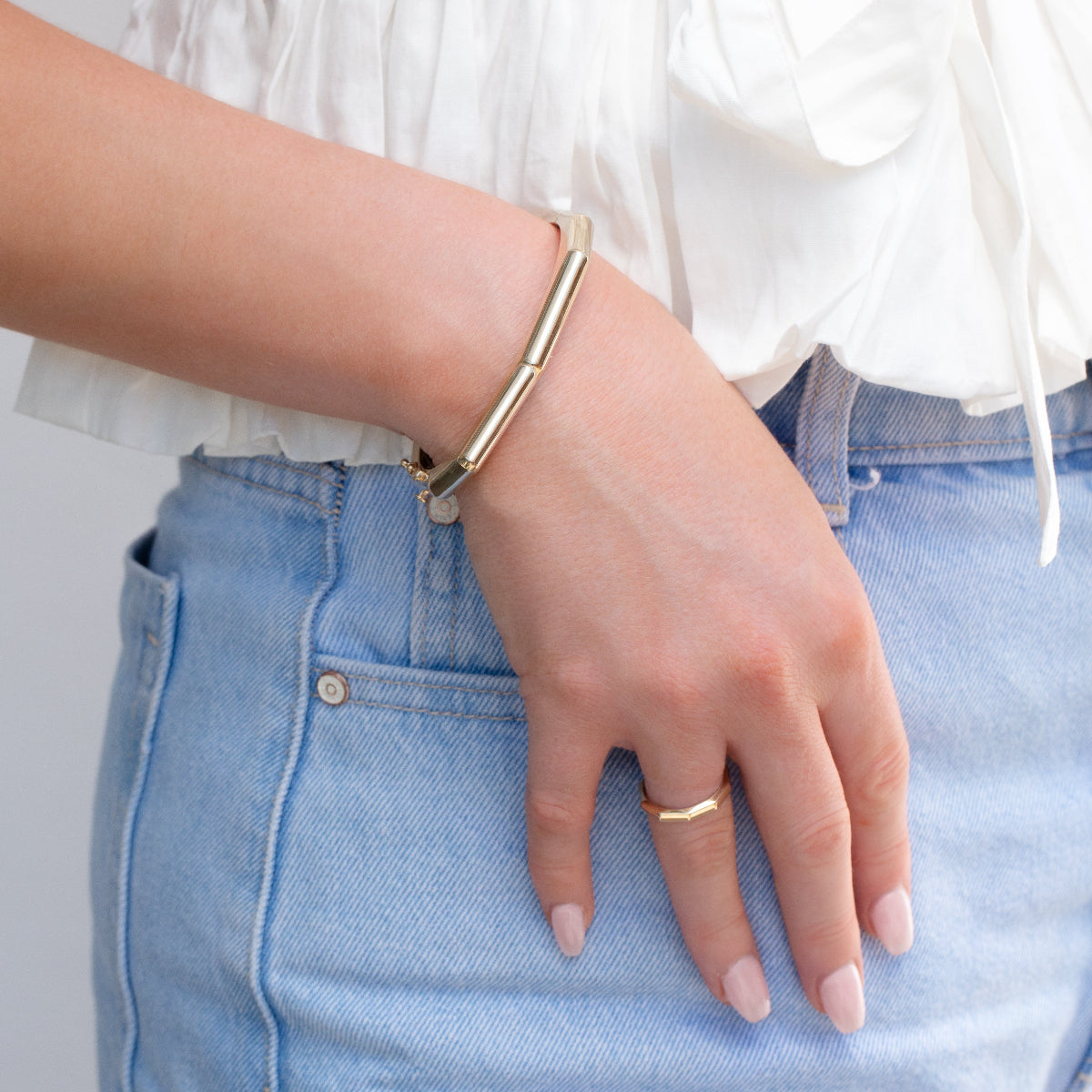 A hand wearing a Tenfold Hinge Bangle and a Tenfold Ring with light pink nails, resting on a light blue jean pocket and styled with a white blouse.