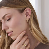 A Link Hoop, Birthstone Hoop, Double Ear Cuff, Paris Ring, Flat Ring and The Peony Bouquet Necklace being worn by a blonde woman with a brown top against a light blurred background. 