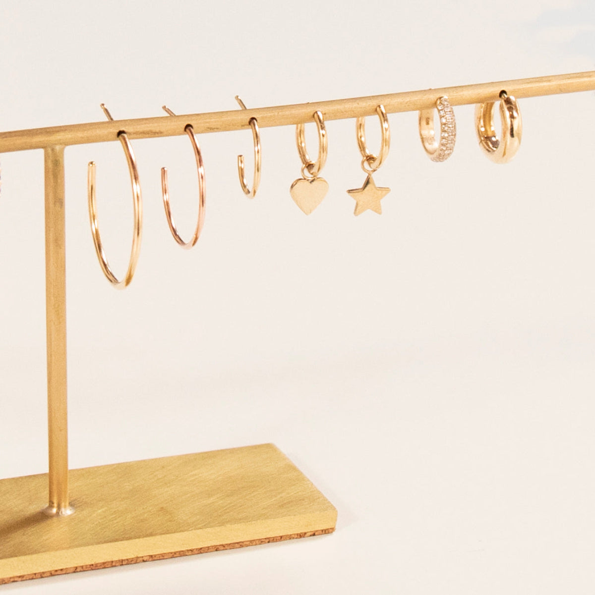 Sleek & Chic Hoops in Large, Medium and Small, Charmed I'm Sure Hoops in Star and Heart, a Pave Huggie and a Gold Huggie, all in yellow gold, displayed on a gold jewellery stand against a light background. 