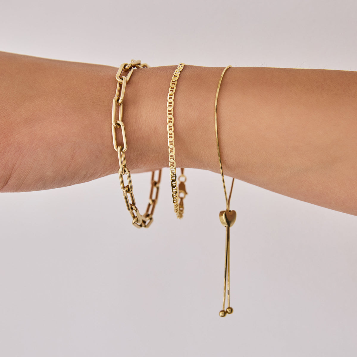 A close-up of an arm wearing The Slide Heart Bracelet, a Bold Link Bracelet and an Overboard Bracelet, all in yellow gold against a light grey background.