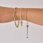 A close-up of an arm wearing The Slide Heart Bracelet, a Bold Link Bracelet and an Overboard Bracelet, all in yellow gold against a light grey background.