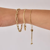 A close-up of an arm wearing The Slide Heart Bracelet, a Bold Link Bracelet and an Overboard Bracelet, all in yellow gold against a light grey background.
