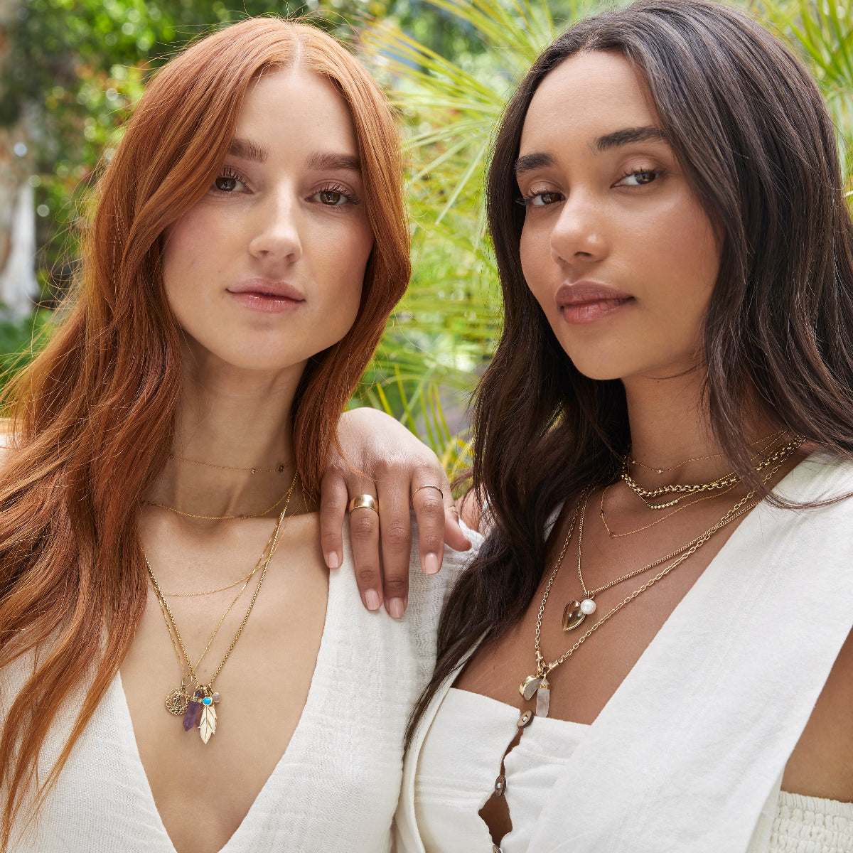 Two women, one wearing Custom Gold Necklaces, The Slide Chain with a Zodiac Pendant, a Box Chain with a Balance Crystal Charm, Turquoise Dangle Charm, Opal Dangle Charm and 20th Anniversary Leaf Pendant, and the other wearing Custom Gold Necklaces, a Rolo Necklace, Diamond Arc Layering Necklace, a Round Curb Chain with a Perla Pendant and Diamond Heart Locket Charm, and an Oval Link Chain with Balance Crystal Charm and Moonstone Crescent Charm, both in white tops, against a blurred leafy background.
