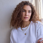 A woman wearing the Ultimate Diamond Necklace, a Bold Link Chain, a Sequence Chain, and a Heart Necklace, all in yellow gold, with a white t-shirt and brown curly hair against a neutral background.