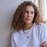 A woman wearing the Ultimate Diamond Necklace, a Bold Link Chain, a Sequence Chain, and a Heart Necklace, all in yellow gold, with a white t-shirt and brown curly hair against a neutral background.