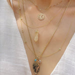 A close-up of a neck wearing a Zodiac Necklace in Gemini, The Happiness Retreat Necklace, a Monogram Necklace with Diamond Yellow Gold with the letter K, Custom Gold Necklace - 3 Letter with the letters S, H and a cross, a Box Chain with a 20th Anniversary Leaf Pendant, a Turquoise Dangle Charm and an Opal Dangle Charm, all in yellow gold, styled with a white linen top. 