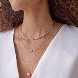Close-up of a woman's décolletage wearing a yellow gold Bold Link Chain, a Pavé MOM Necklace and a Perla Necklace, with brown curly hair and a white top.