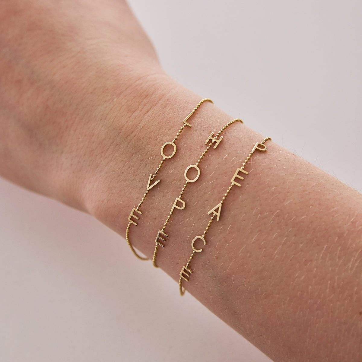 A wrist wearing two Custom Gold Bracelets – 4 Letters (L, O, V, E and H, O, P, E) and one 5 Letters (P, E, A, C, E) in bead chains, on a neutral background.