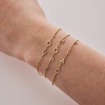 A wrist wearing two Custom Gold Bracelets – 4 Letters (L, O, V, E and H, O, P, E) and one 5 Letters (P, E, A, C, E) in bead chains, on a neutral background.