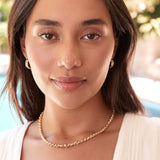 A brunette woman wearing a yellow gold Rolo Necklace, Link Hoops and a white top with a blurred outdoor background. 