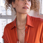 A woman wearing a yellow gold Bold Link Chain, an Element Short Link Chain with a Pink Opal Tear Drop Charm, and a Pavé Charm Stud - Paw, with an orange shirt and brown curly hair against a blurred indoor background.