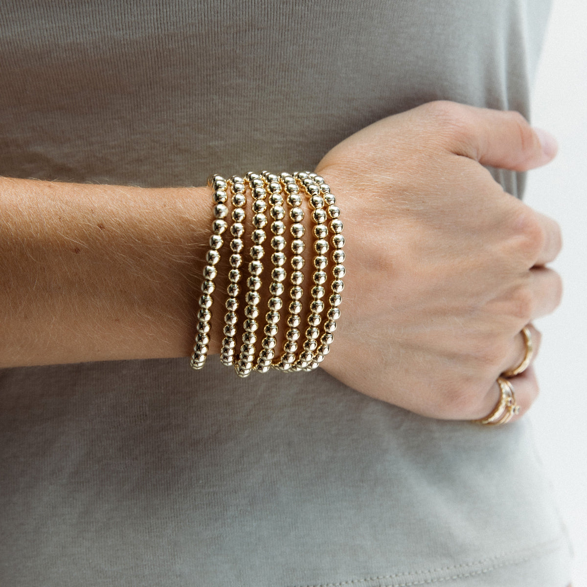 A close up a person's wrist wearing multiple stacked Luxe Bubble Bracelets and gold rings, with a sage green top. 