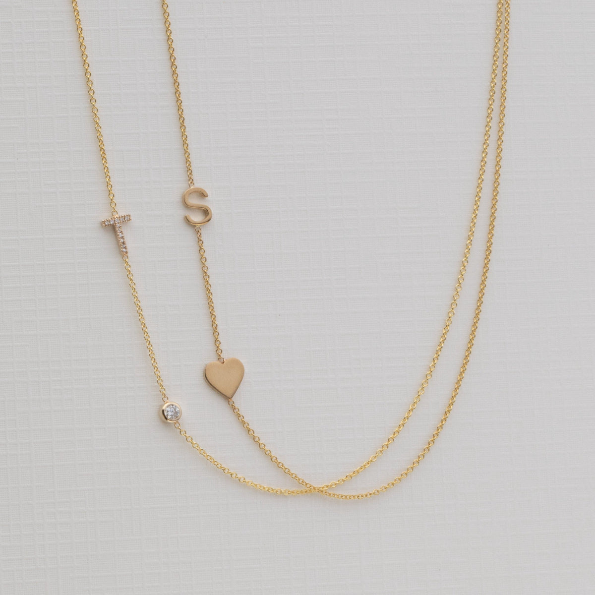 A Monogram Necklace with Heart Yellow Gold and a letter S charm, and a Pavé Monogram Necklace with Diamond and a letter T charm against an off-white background. 