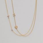 A Monogram Necklace with Heart Yellow Gold and a letter S charm, and a Pavé Monogram Necklace with Diamond and a letter T charm against an off-white background. 