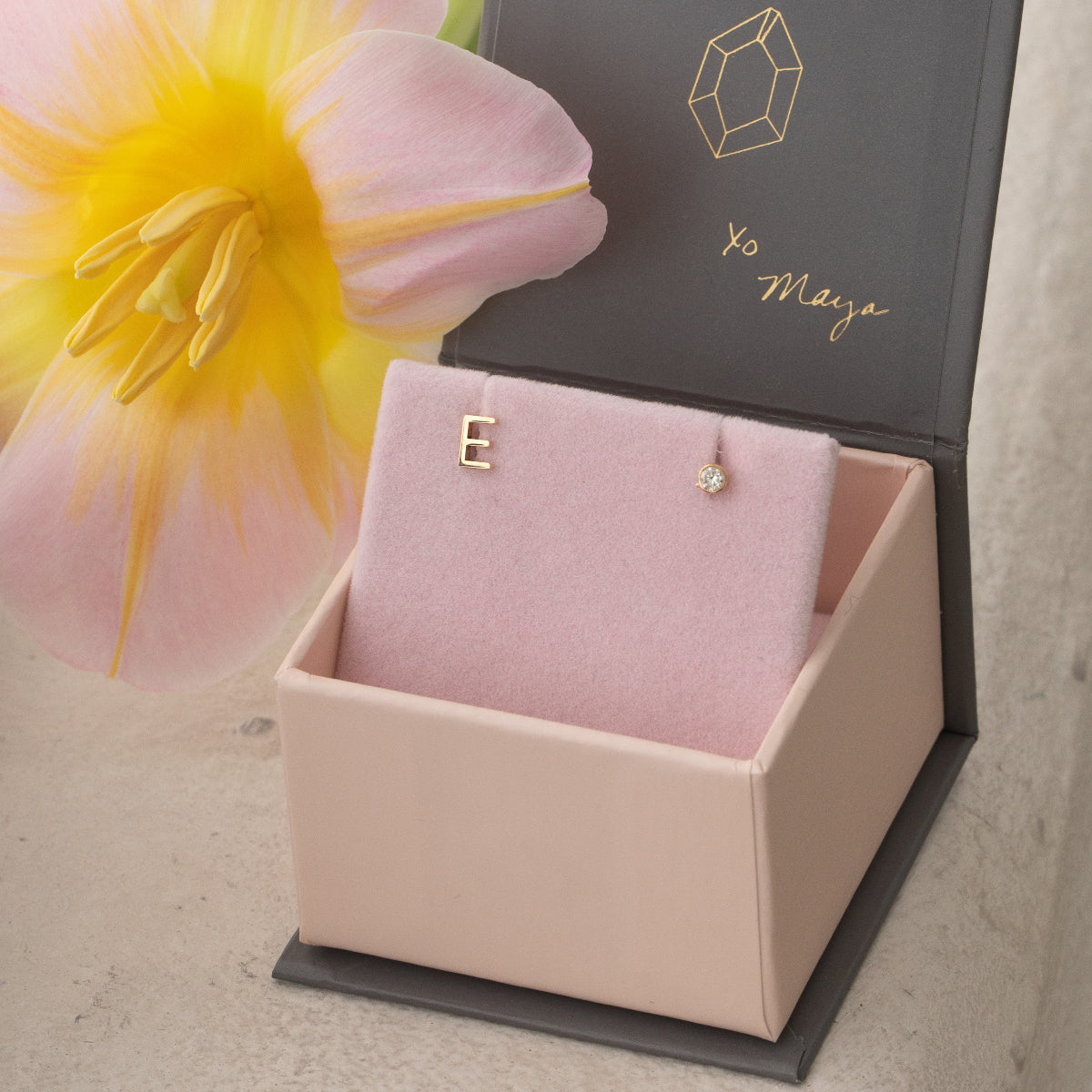 A yellow gold monogram earring set featuring a mini letter E stud and a mini diamond stud, placed in a Maya Brenner pink earring case with a yellow and pink flower.