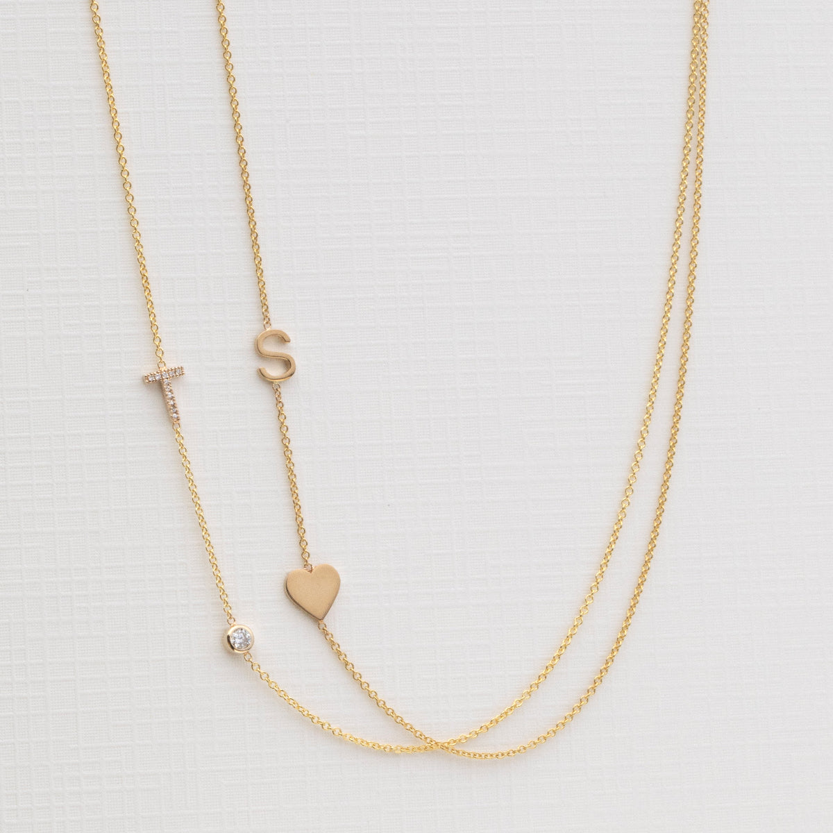 A Monogram Necklace with Heart Yellow Gold and a letter S charm, and a Pavé Monogram Necklace with Diamond and a letter T charm against an off-white background. 