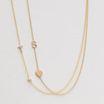 A Monogram Necklace with Heart Yellow Gold and a letter S charm, and a Pavé Monogram Necklace with Diamond and a letter T charm against an off-white background. 