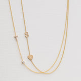 A Monogram Necklace with Heart Yellow Gold and a letter S charm, and a Pavé Monogram Necklace with Diamond and a letter T charm against an off-white background. 