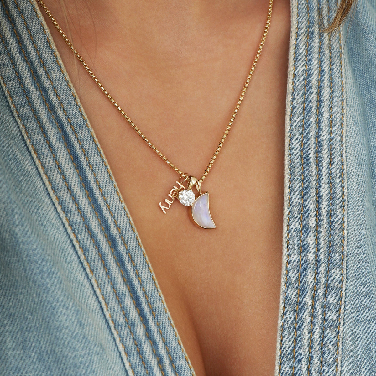 Close-up of a neck wearing a yellow gold Box Chain with a Petite Nameplate Pendant with 'henry', a Queen Diamond Pendant and a Moonstone Crescent Charm, styled with a light blue denim top. 
