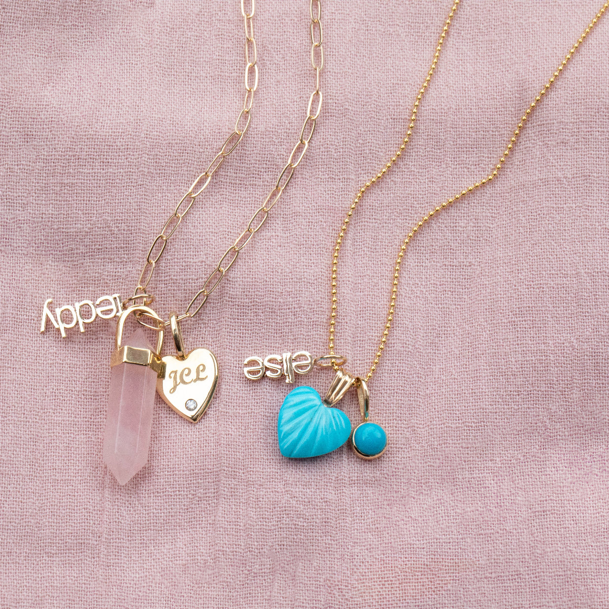 A yellow gold Element Short Link Chain with a Petite Nameplate Pendant in 'teddy', a Rose Quartz Balance Crystal Charm and an Engraved Heart Charm, and a yellow gold Classic Bead Chain with a Petite Nameplate Pendant in 'elise', an Etched Turquoise Heart Charm and a Turquoise Dangle Charm, against a light pink fabric background.