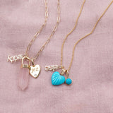 A yellow gold Element Short Link Chain with a Petite Nameplate Pendant in 'teddy', a Rose Quartz Balance Crystal Charm and an Engraved Heart Charm, and a yellow gold Classic Bead Chain with a Petite Nameplate Pendant in 'elise', an Etched Turquoise Heart Charm and a Turquoise Dangle Charm, against a light pink fabric background.