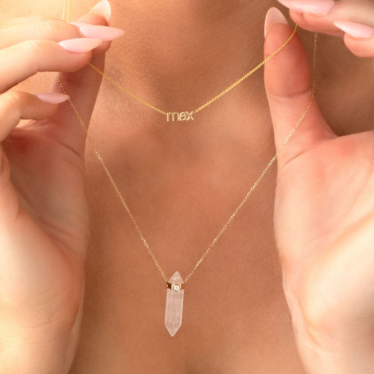 A close-up of a neck wearing a yellow gold Petite Name Plate Necklace with “max” and the Clarity Retreat Necklace, with hands holding the necklaces.