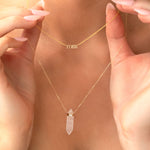 A close-up of a neck wearing a yellow gold Petite Name Plate Necklace with “max” and the Clarity Retreat Necklace, with hands holding the necklaces.