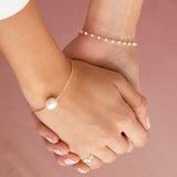 Two hands holding each other wearing The Lily Bouquet Bracelet, a Baroque White Pearl Bangle and a Perla Pinky Ring on a pink background. 