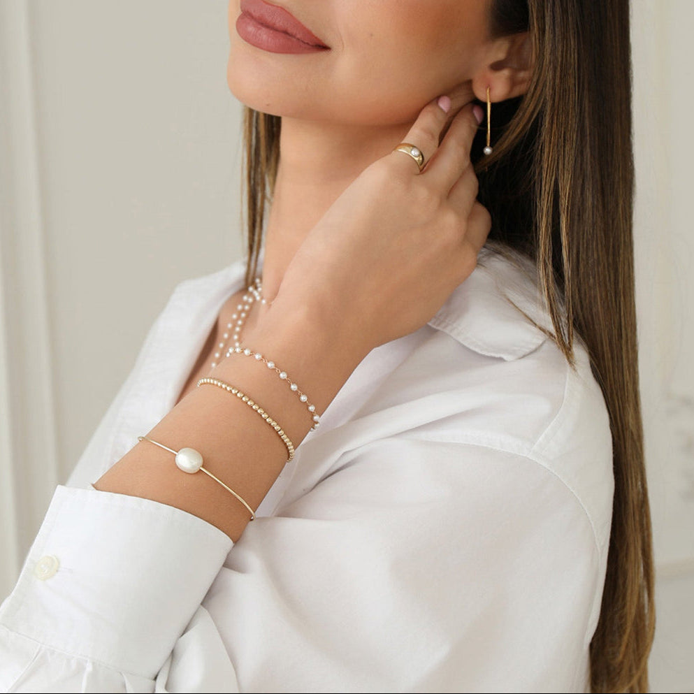 A woman wearing The Lily Bouquet Bracelet, Baroque White Pearl Bangle, Little Bubble Bracelet, Perla Pinky Ring, Pearl Drift Threader and The Lily Bouquet Necklace, styled with a white shirt. 