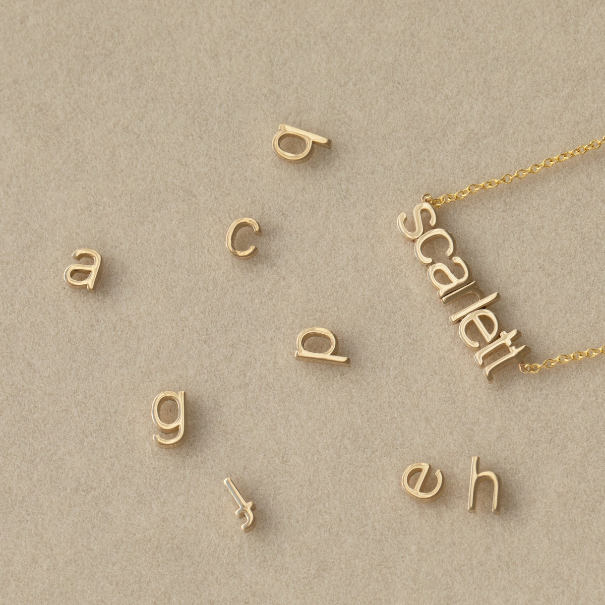A Petite Nameplate Necklace in Scarlett with several Petite Letter Charms, all in yellow gold, against a light beige background. 